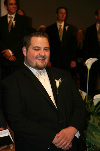 Smiling man seated, facing the camera, wearing a black tux
